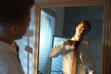 Photo of Handsome man adjusting necktie while getting ready near mirror indoors
