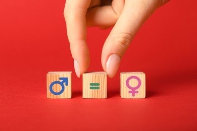 Image of Sex education. Woman putting cube with equals sign between cubes with female and male gender symbols on red background, closeup