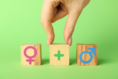 Image of Sex education. Woman putting cube with plus sign between cubes with female and male gender symbols on green background, closeup