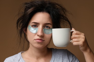 Photo of Woman with eye patches and cup of drink suffering from hangover on brown background