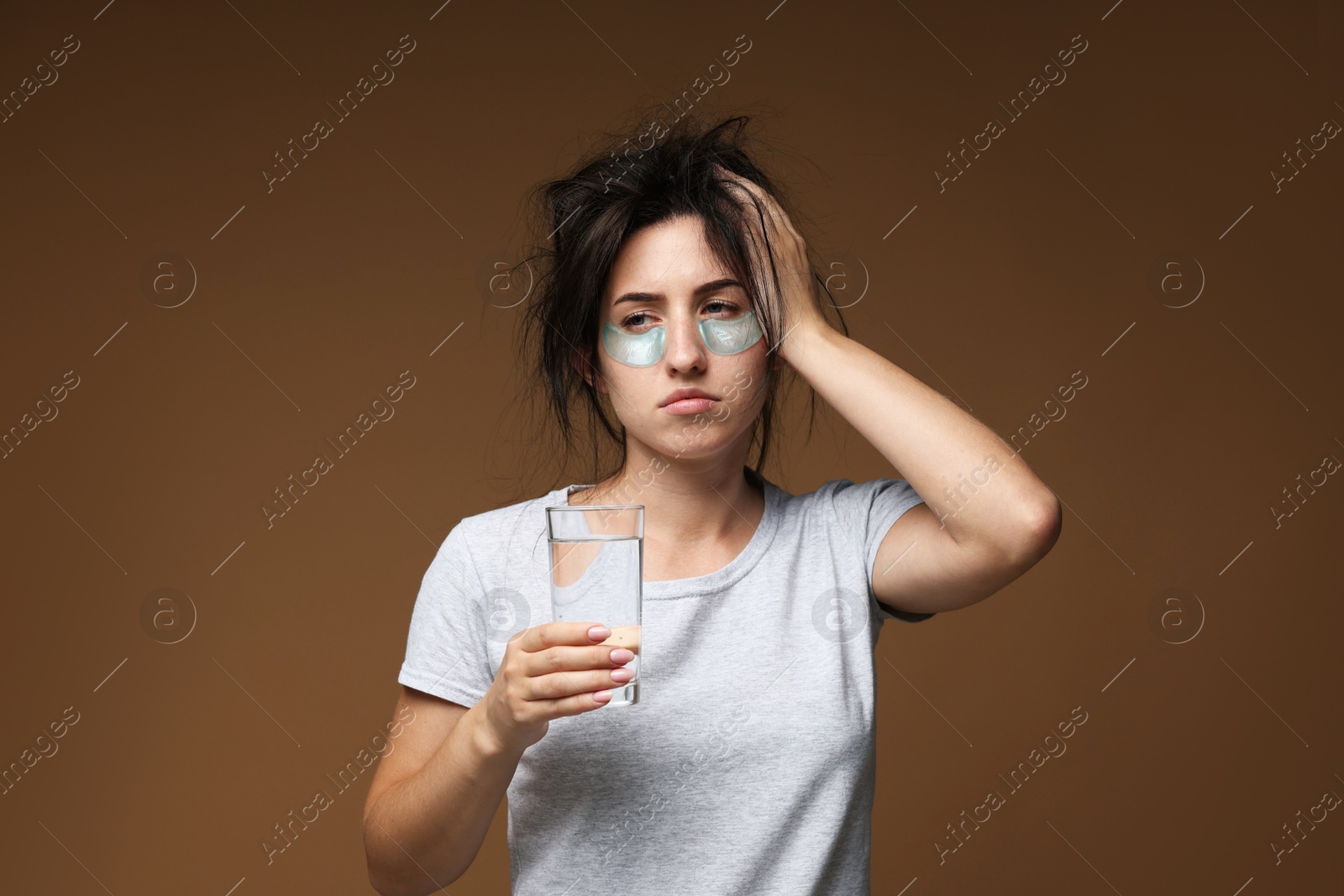 Photo of Woman with eye patches and glass of water suffering from hangover on brown background