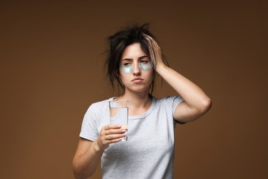 Photo of Woman with eye patches and glass of water suffering from hangover on brown background