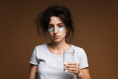 Photo of Woman with eye patches and glass of water suffering from hangover on brown background