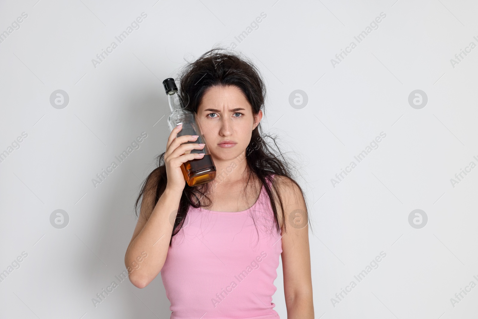 Photo of Woman with bottle of whiskey suffering from hangover on light grey background