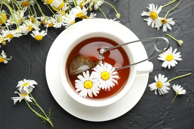 Photo of Tasty chamomile tea, infuser and flowers on black table, flat lay