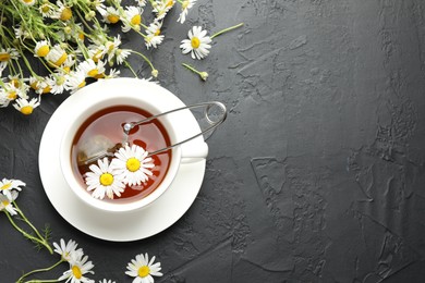 Photo of Tasty chamomile tea, infuser and flowers on black table, flat lay. Space for text