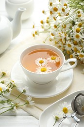 Photo of Tasty chamomile tea, infuser and flowers on white wooden table, closeup