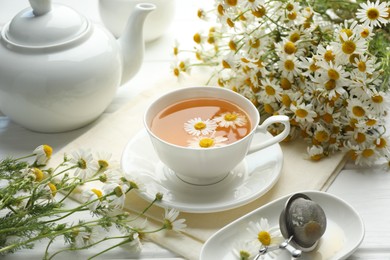 Photo of Tasty chamomile tea, infuser and flowers on white wooden table, closeup