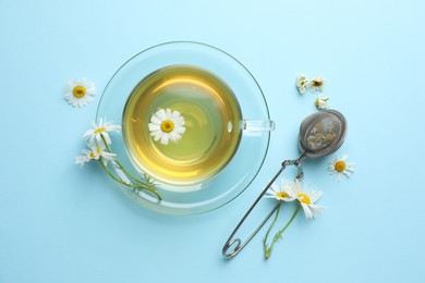 Photo of Tasty chamomile tea, infuser and flowers on light blue background, flat lay