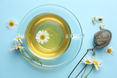 Photo of Tasty chamomile tea, infuser and flowers on light blue background, flat lay