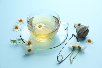 Photo of Tasty chamomile tea, infuser and flowers on light blue background, closeup