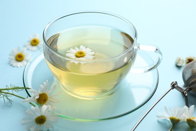 Photo of Tasty chamomile tea, infuser and flowers on light blue background, closeup