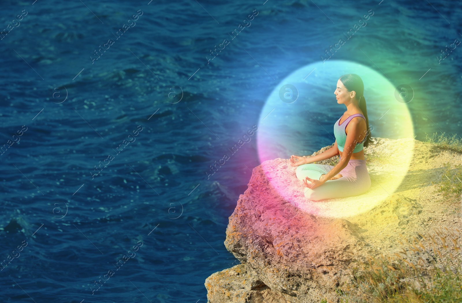 Image of Woman meditating in Lotus pose on cliff near sea. Rainbow bubble around her representing aura
