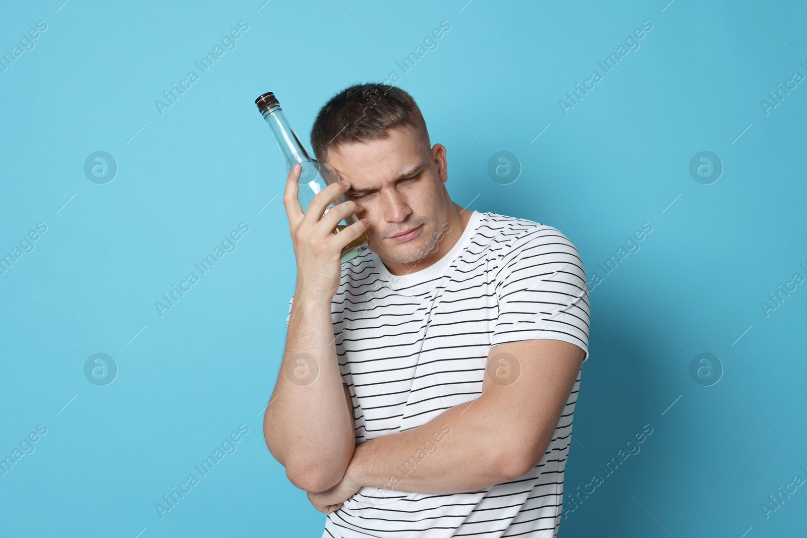 Photo of Man with bottle of whiskey suffering from hangover on light blue background