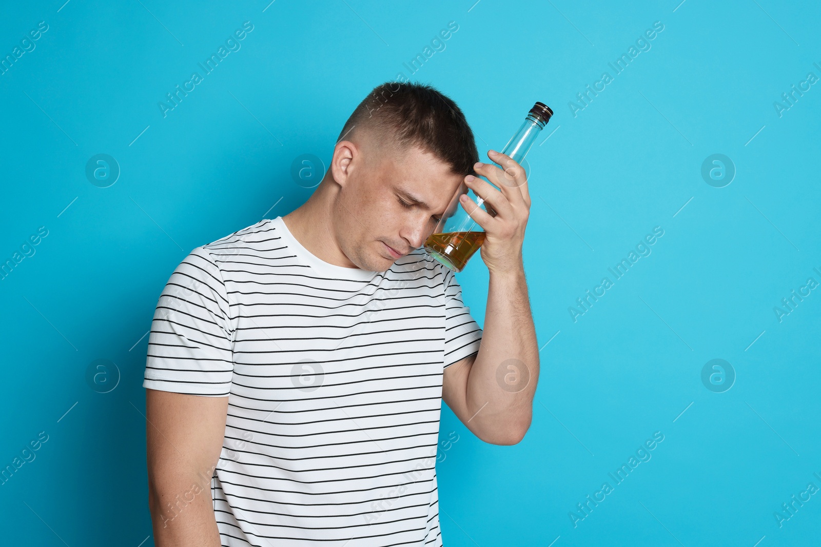 Photo of Man with bottle of whiskey suffering from hangover on light blue background