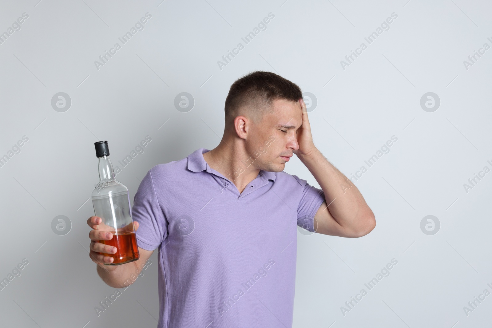 Photo of Man with bottle of whiskey suffering from hangover on light grey background