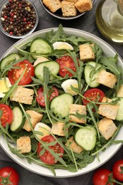 Photo of Tasty salad with croutons, cucumbers, tomatoes and arugula on black table, flat lay