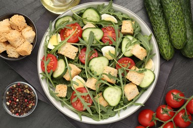 Photo of Tasty salad with croutons, cucumbers, tomatoes and arugula on black table, flat lay