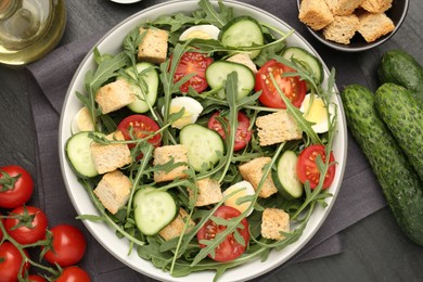 Photo of Tasty salad with croutons, cucumbers, tomatoes and arugula on black table, flat lay