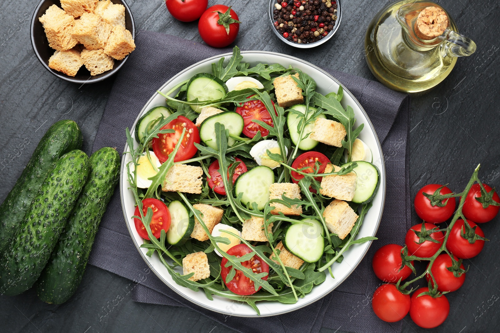 Photo of Tasty salad with croutons, cucumbers, tomatoes and arugula on black table, flat lay