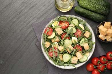 Photo of Tasty salad with croutons, cucumbers, tomatoes and arugula on black table, flat lay. Space for text
