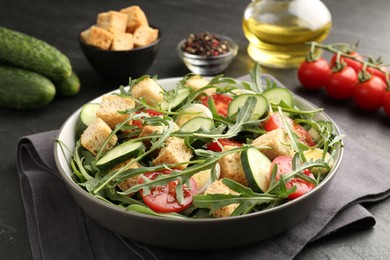 Photo of Tasty salad with croutons, cucumbers, tomatoes and arugula on black table, closeup