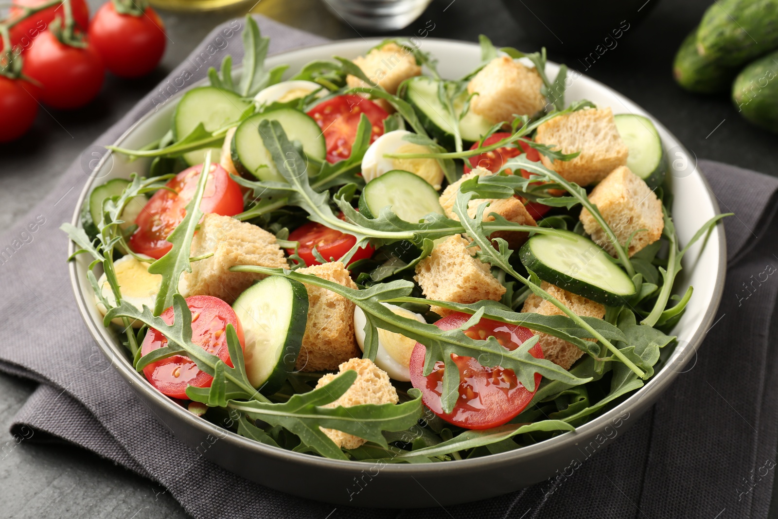Photo of Tasty salad with croutons, cucumbers, tomatoes and arugula on black table, closeup