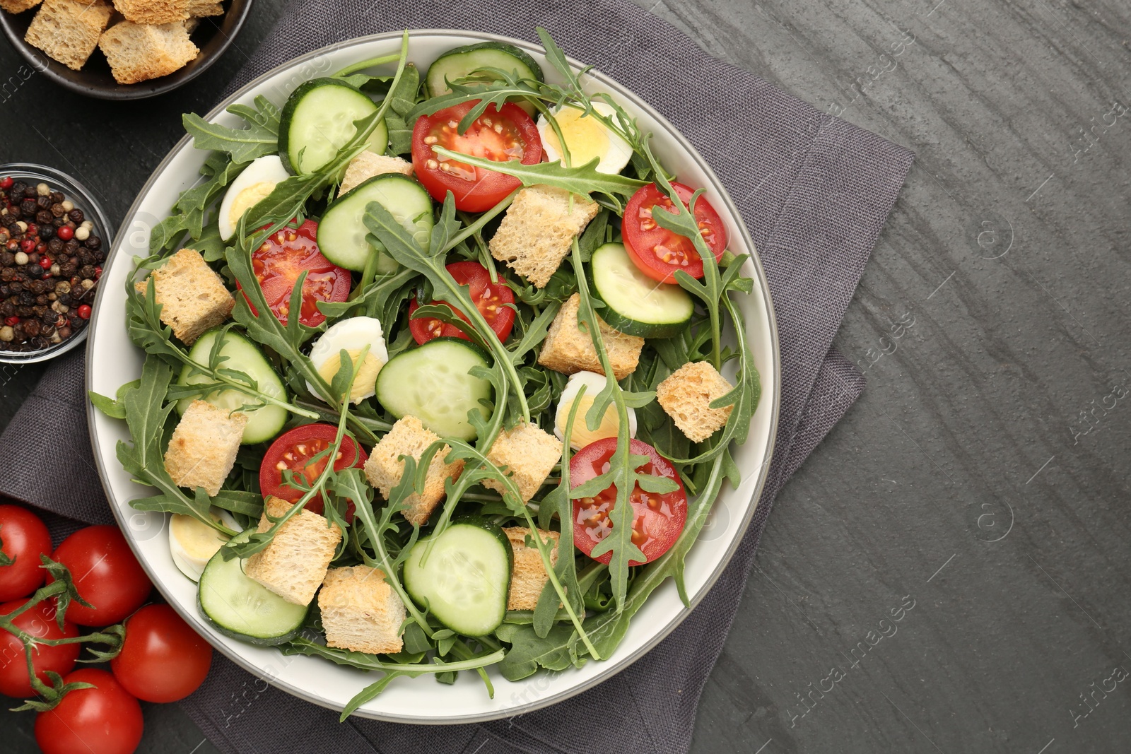 Photo of Tasty salad with croutons, cucumbers, tomatoes and arugula on black table, flat lay. Space for text