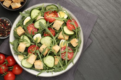 Photo of Tasty salad with croutons, cucumbers, tomatoes and arugula on black table, flat lay. Space for text