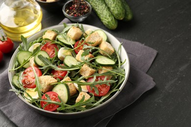 Photo of Tasty salad with croutons, cucumbers, tomatoes and arugula on black table, closeup