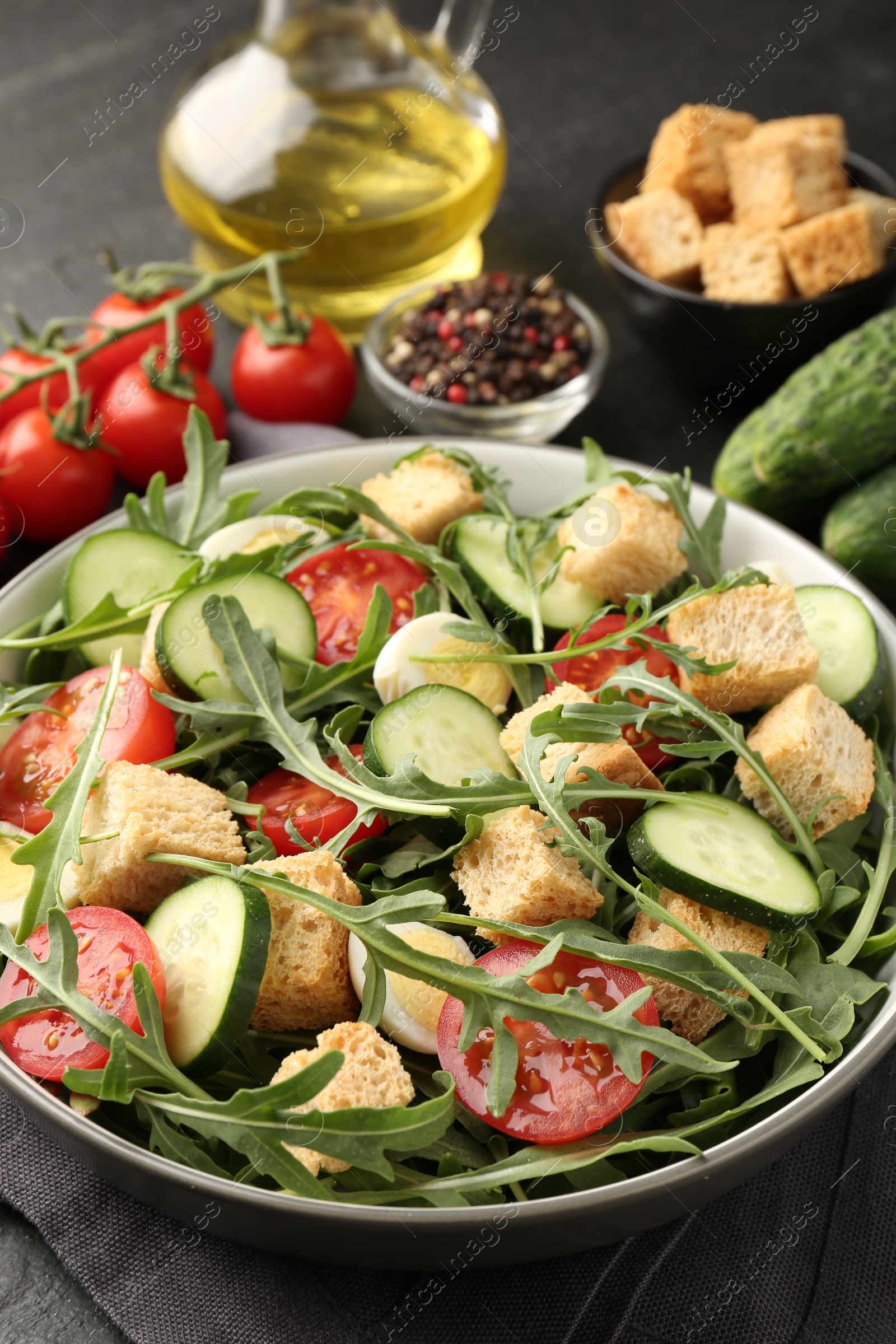 Photo of Tasty salad with croutons, cucumbers, tomatoes and arugula on black table, closeup