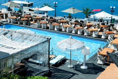 Photo of Beautiful swimming pool, sunbeds, tables, chairs and umbrellas near sea on sunny day