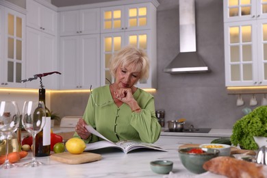Photo of Senior woman with glasses reading magazine at marble table in kitchen