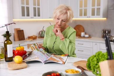 Photo of Senior woman with glasses reading magazine at marble table in kitchen