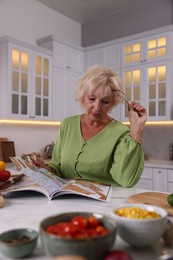 Photo of Senior woman with glasses reading magazine at marble table in kitchen