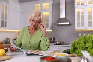 Photo of Senior woman in glasses reading magazine at marble table in kitchen