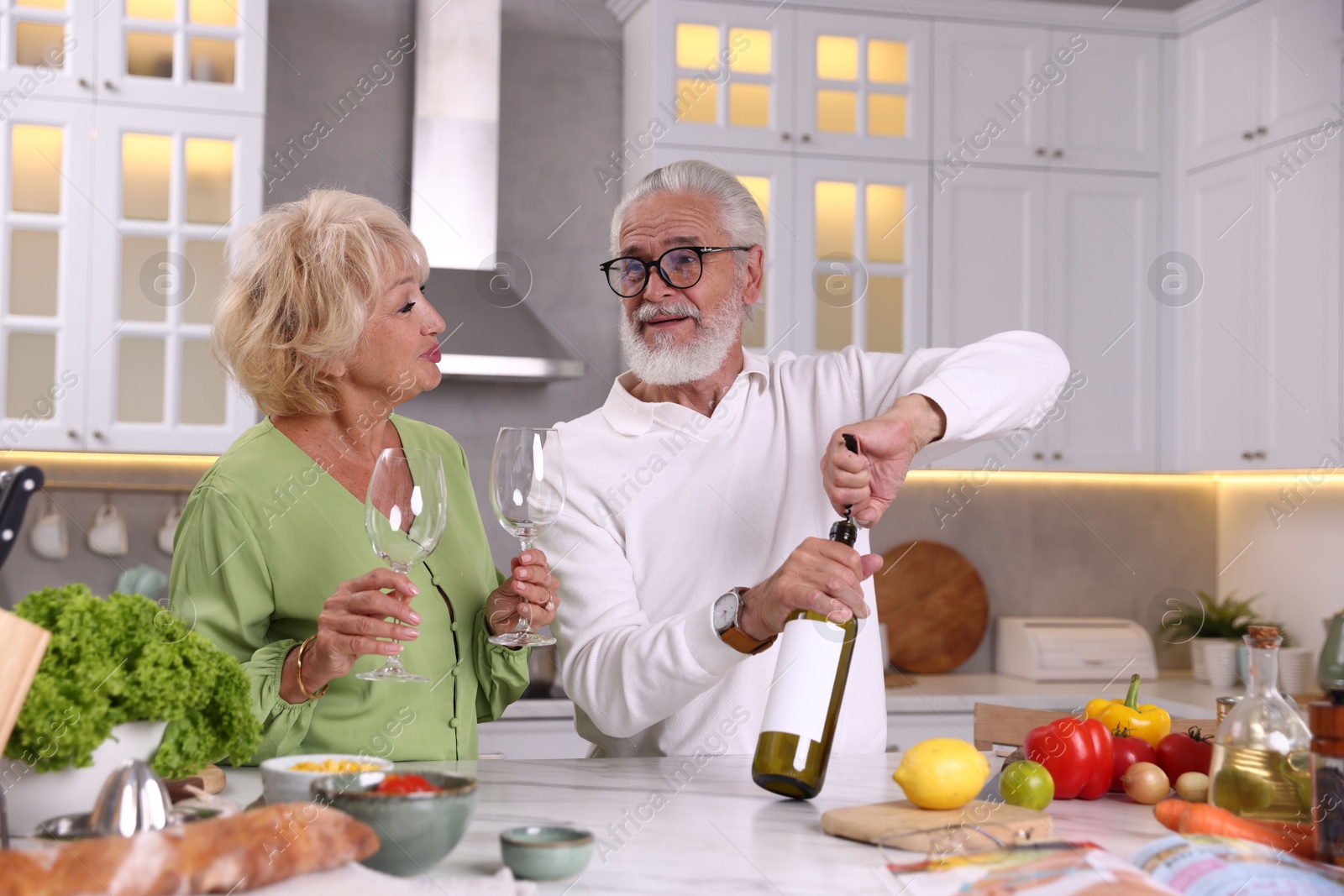 Elderly man opening bottle of wine while cooking with his wife at table in kitchen Photo of Elderly man opening bottle of wine while cooking with his wife at table in kitchen