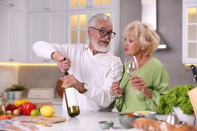Photo of Elderly man opening bottle of wine while cooking with his wife at table in kitchen