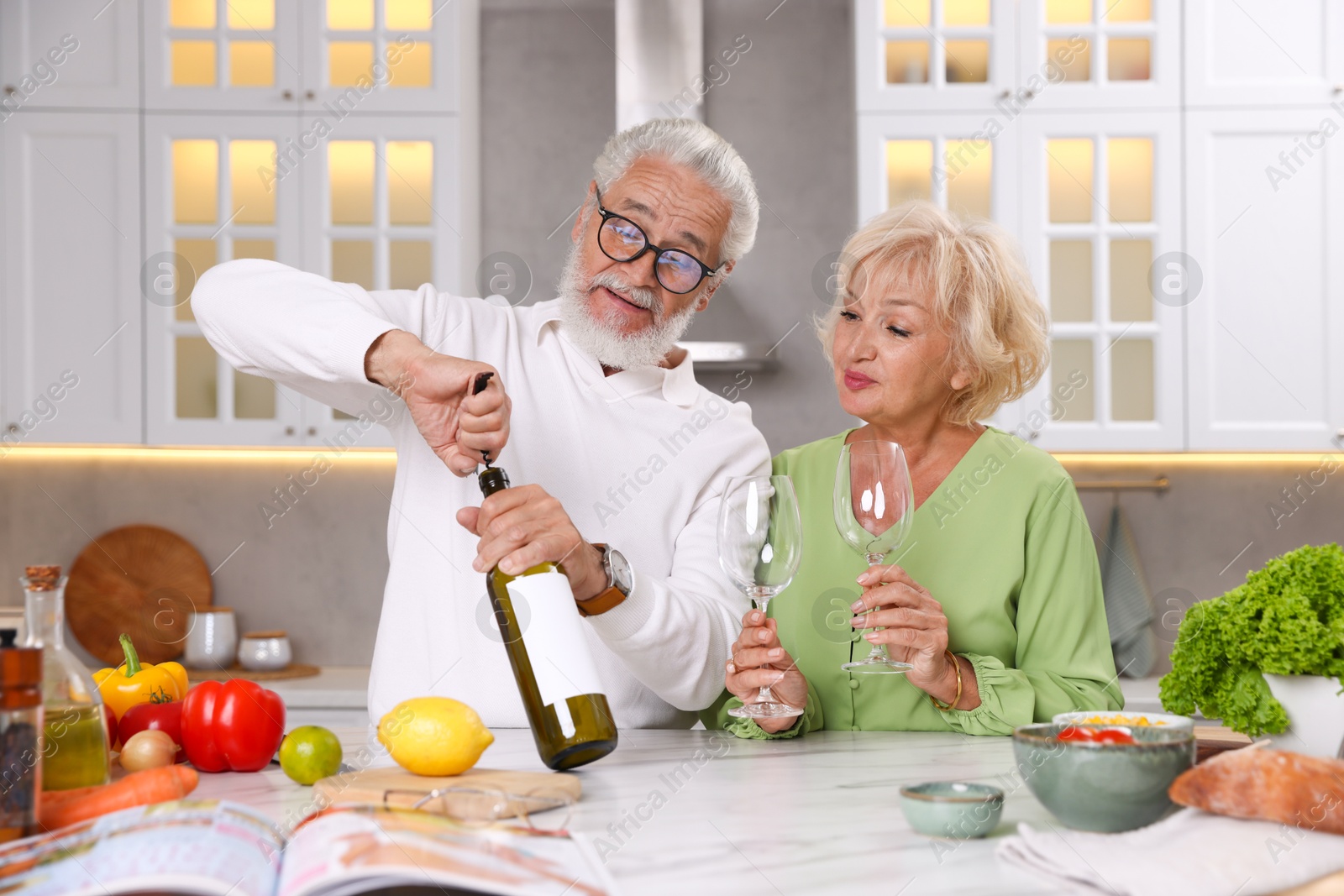 Elderly man opening bottle of wine while cooking with his wife at table in kitchen Photo of Elderly man opening bottle of wine while cooking with his wife at table in kitchen