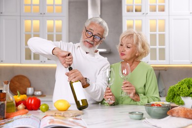 Photo of Elderly man opening bottle of wine while cooking with his wife at table in kitchen