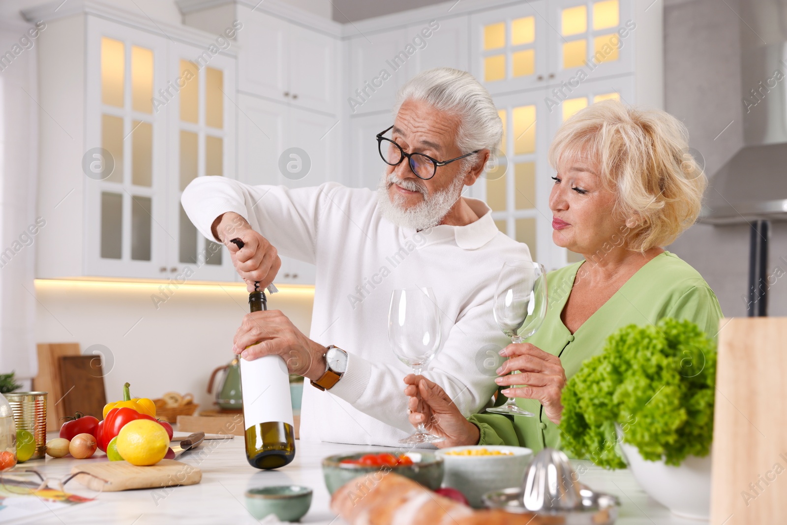 Elderly man opening bottle of wine while cooking with his wife at table in kitchen Photo of Elderly man opening bottle of wine while cooking with his wife at table in kitchen