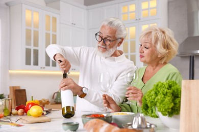 Photo of Elderly man opening bottle of wine while cooking with his wife at table in kitchen