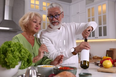 Photo of Elderly man opening bottle of wine while cooking with his wife at table in kitchen