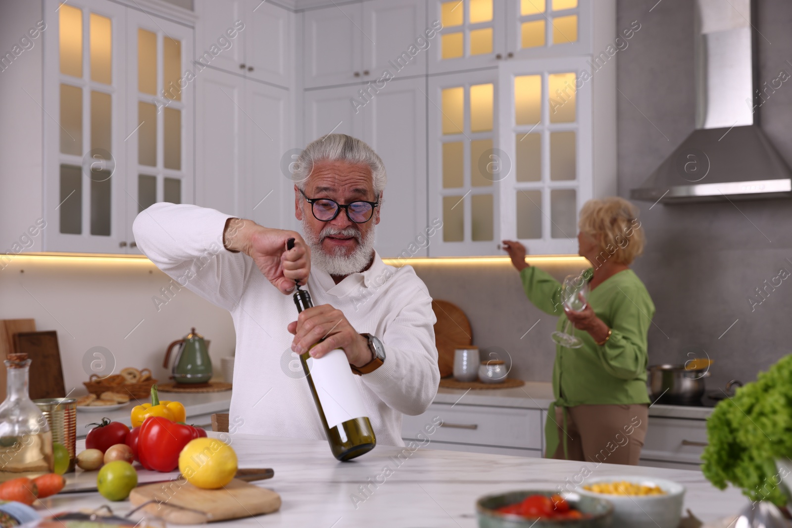 Elderly man opening bottle of wine while cooking with his wife at table in kitchen, selective focus Photo of Elderly man opening bottle of wine while cooking with his wife at table in kitchen, selective focus