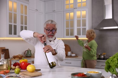 Photo of Elderly man opening bottle of wine while cooking with his wife at table in kitchen, selective focus