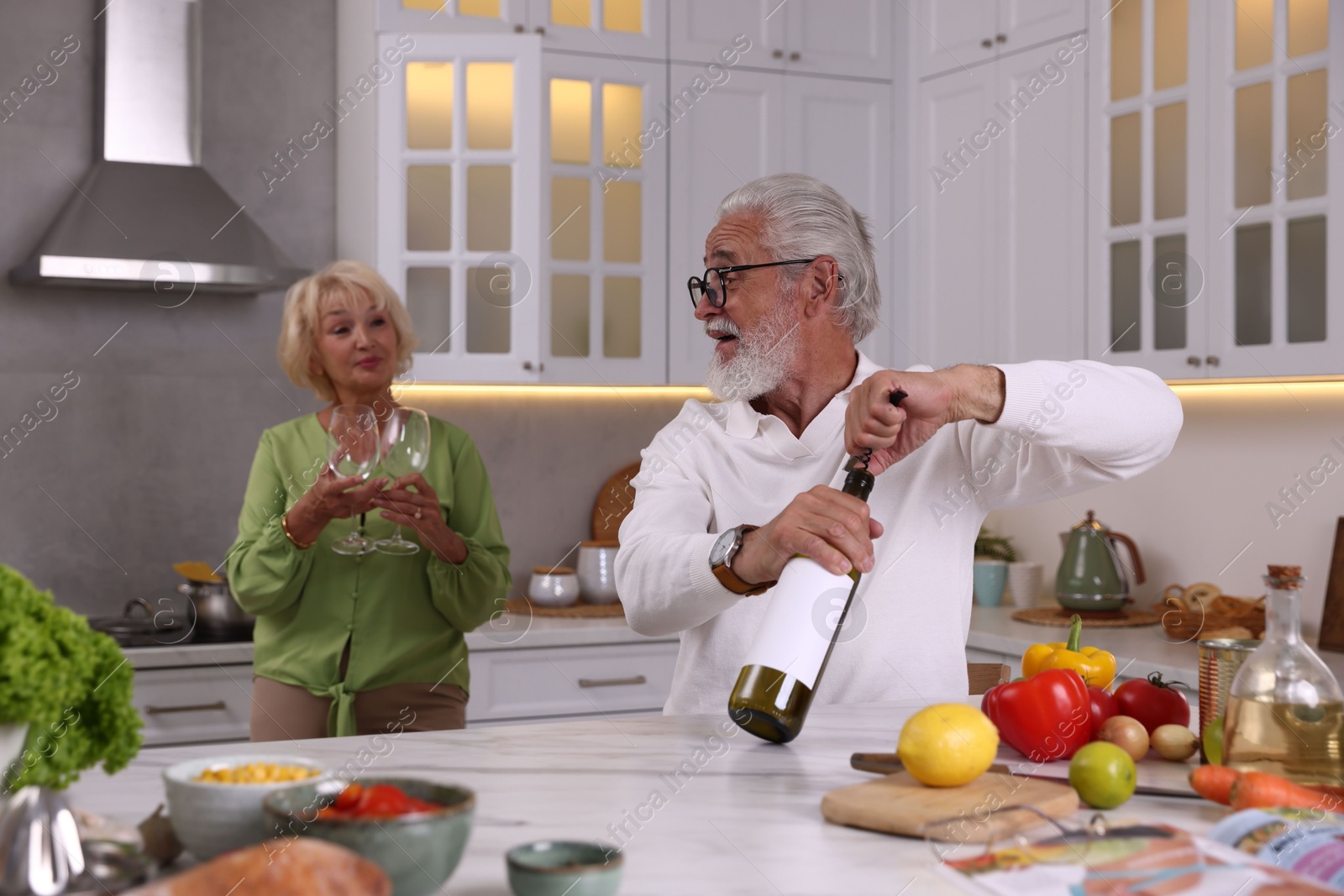 Elderly man opening bottle of wine while cooking with his wife at table in kitchen, selective focus Photo of Elderly man opening bottle of wine while cooking with his wife at table in kitchen, selective focus