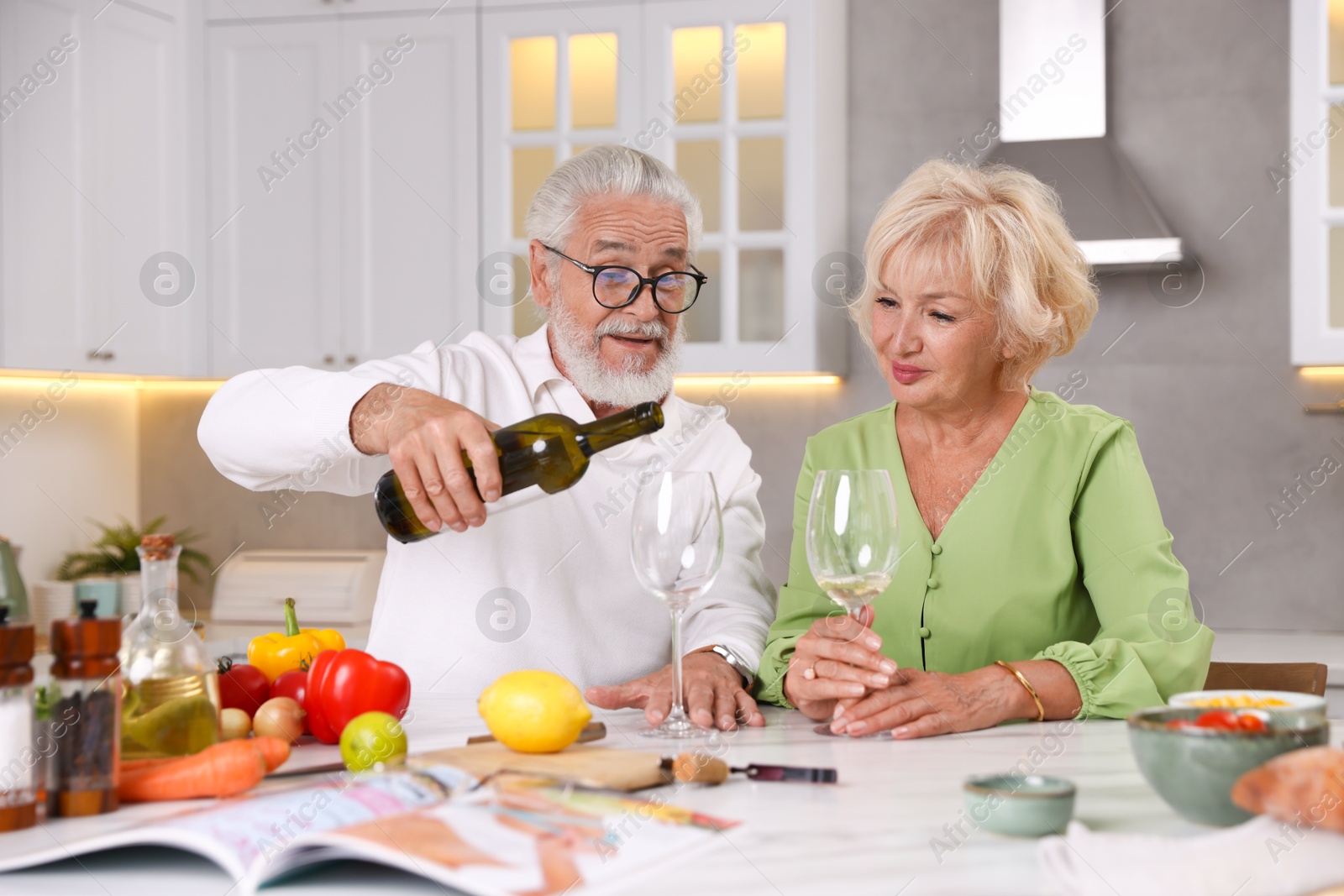 Elderly couple drinking white wine while cooking at table in kitchen Photo of Elderly couple drinking white wine while cooking at table in kitchen
