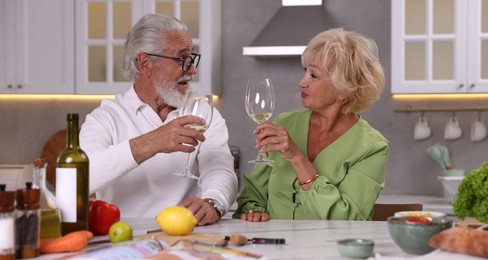 Photo of Elderly couple drinking white wine while cooking at table in kitchen