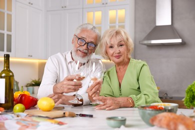 Photo of Elderly couple drinking white wine while cooking at table in kitchen