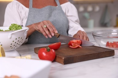 Photo of Senior woman cutting tomato at white marble table in kitchen, closeup